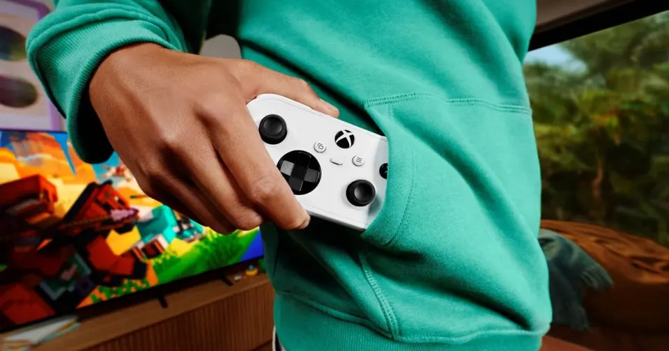 A hand sliding the small white Xbox Nano Controller out of a green hoodie pocket in front of a colorful game on a TV.